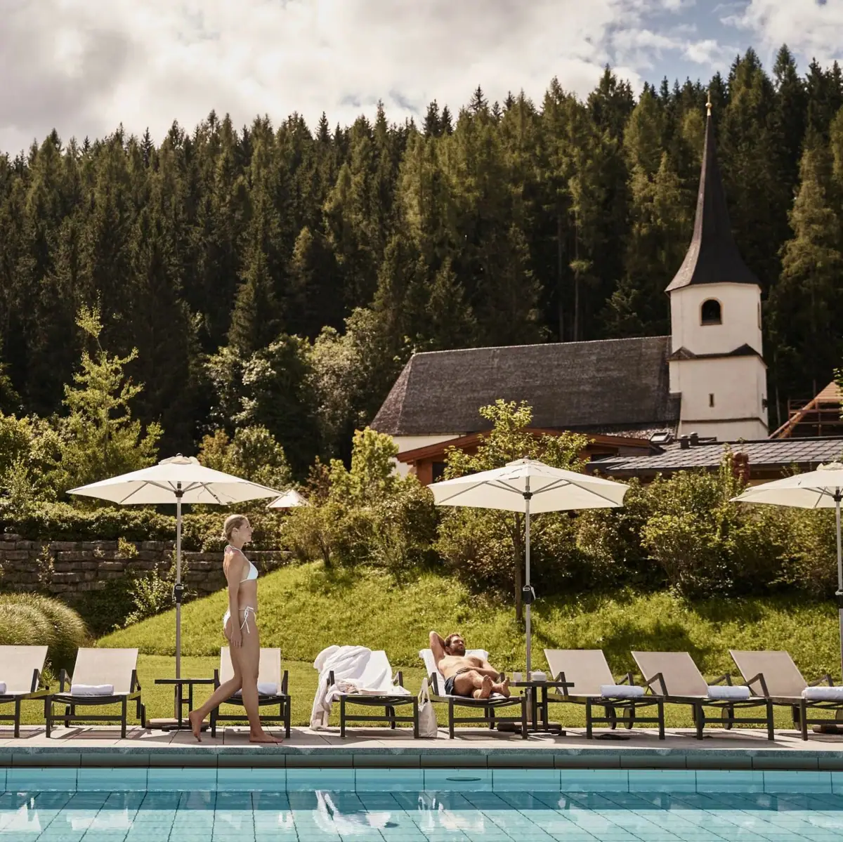 Idyllic pool landscape in Werfenweng with a view of a small church and a dense forest. A woman in a swimming costume strolls along the pool while a guest relaxes on a sun lounger under a parasol.