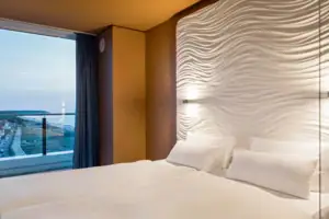 Suite at the aja Warnemünde Bed with white sheets in front of a wall with a window overlooking the sea