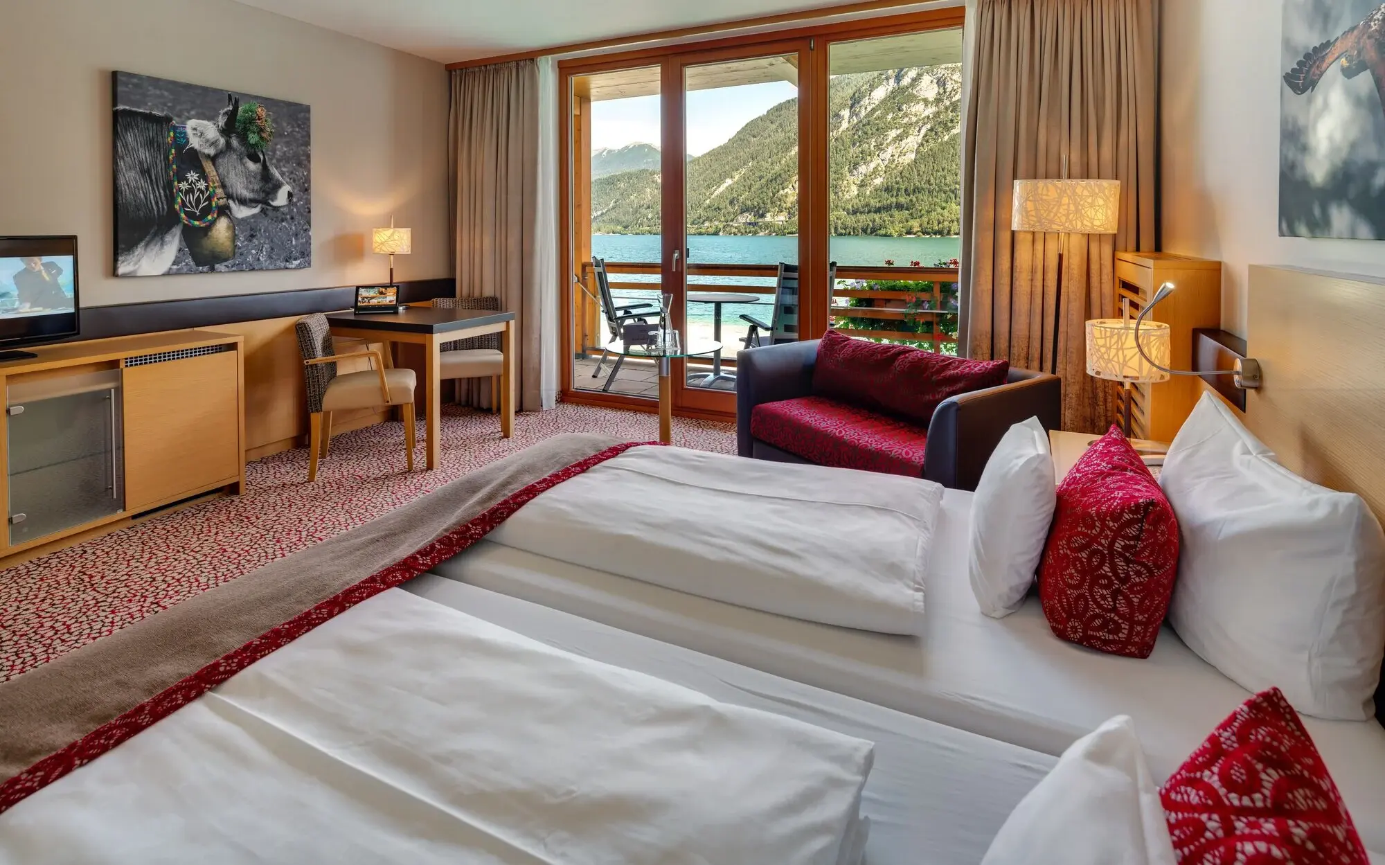 Interior of a bedroom with a bed, a sofa and a table. In the background you can see a balcony with a view of Lake Achensee.