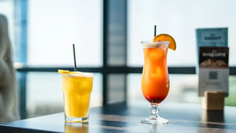 Two glasses of orange cocktails with garnishes and straws on a table.