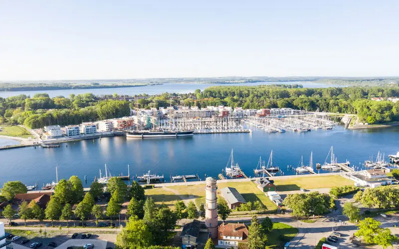 Aerial view of a picturesque marina with numerous sailboats, modern waterfront buildings, and lush green surroundings, set against a calm blue lake under a clear sky.