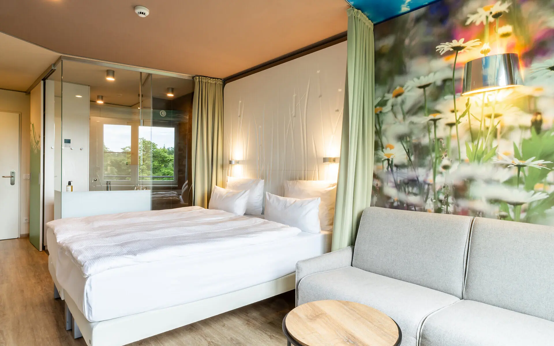 Light hotel room with a comfortable bed, glas bathroom und seating area. The wooden floor and realistic flour wallpaper complement the light and natural flair of the room. 