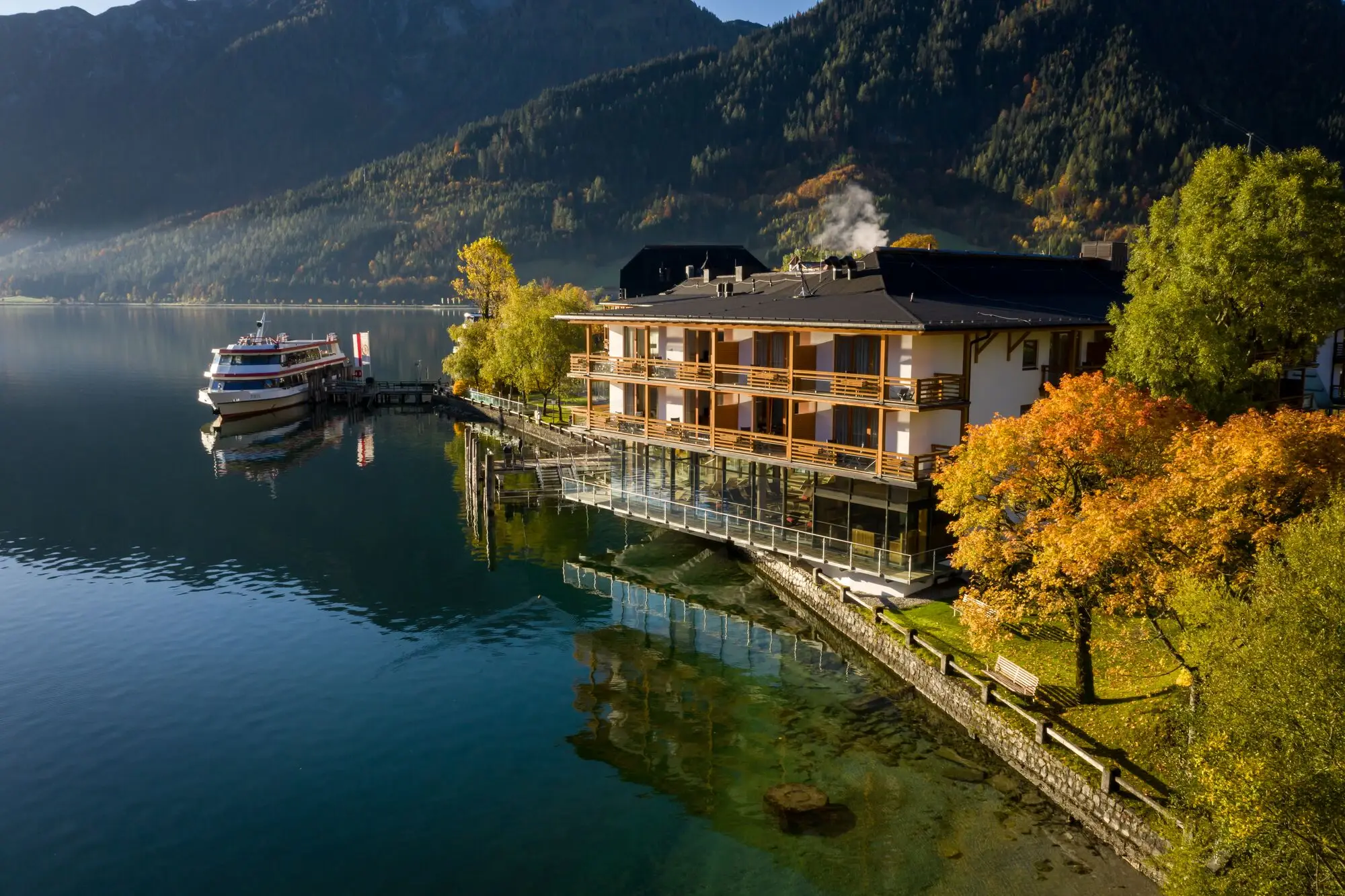 aja Fürstenhaus am Achensee Building by the water with trees and mountains in the background.