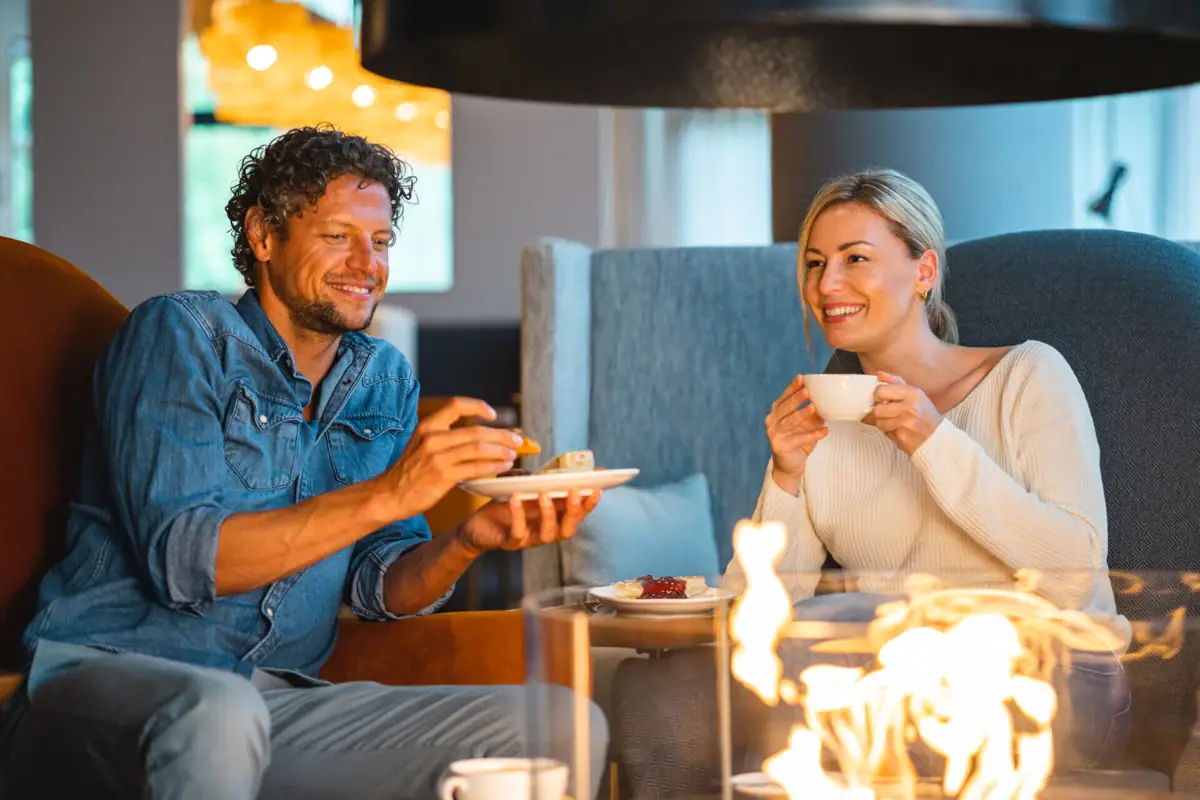 A man and a woman sit on a chair and hold coffee cups by the fireplace with a burning fire.