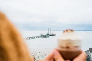 Eine Person hält eine heiße Schokolade mit Sahne und im Hintergrund ist ein Pier zu sehen.