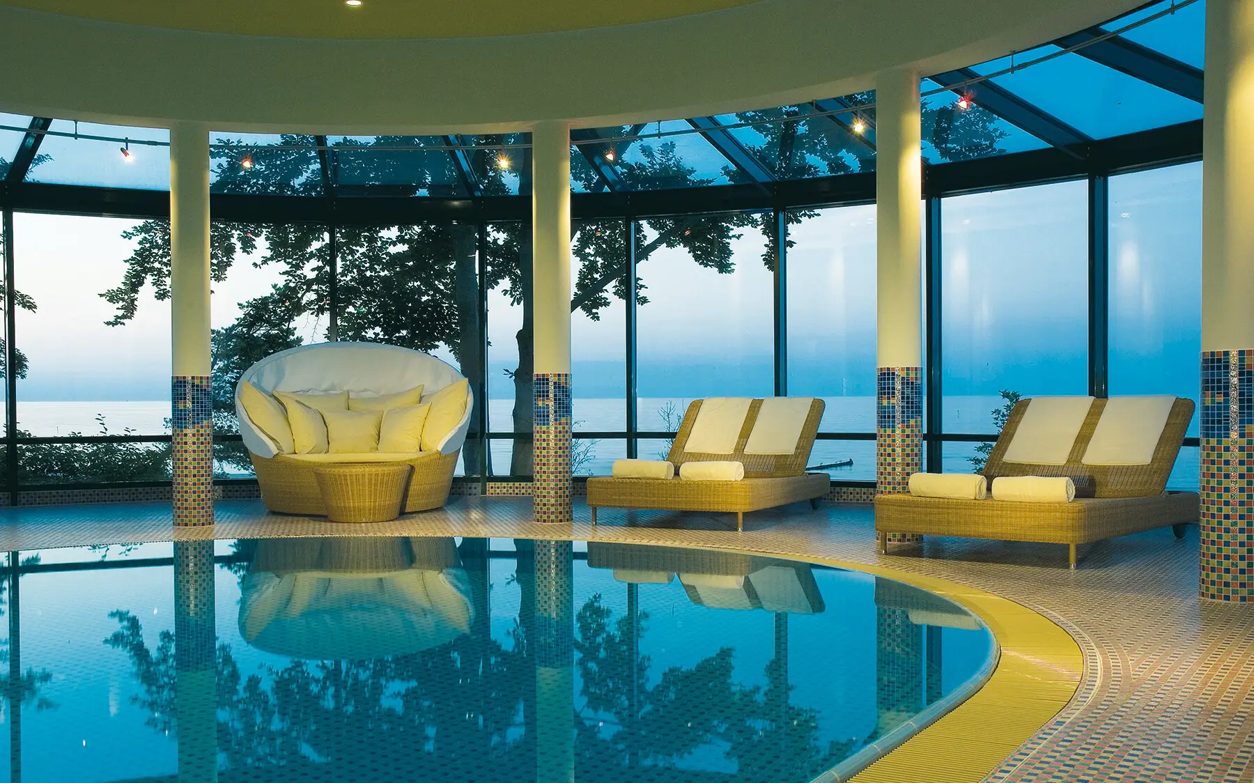 Indoor pool with chairs by a large window overlooking trees.