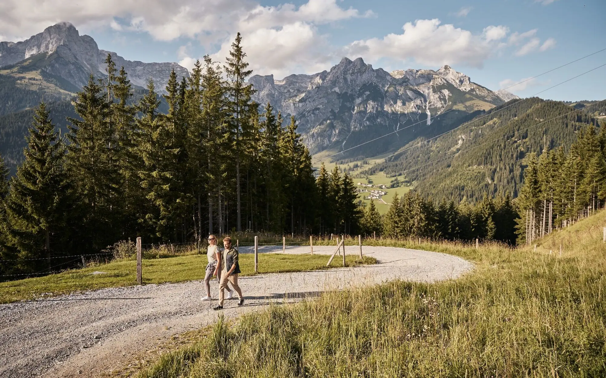 Two people are walking on a gravel path in front of a mountain range.