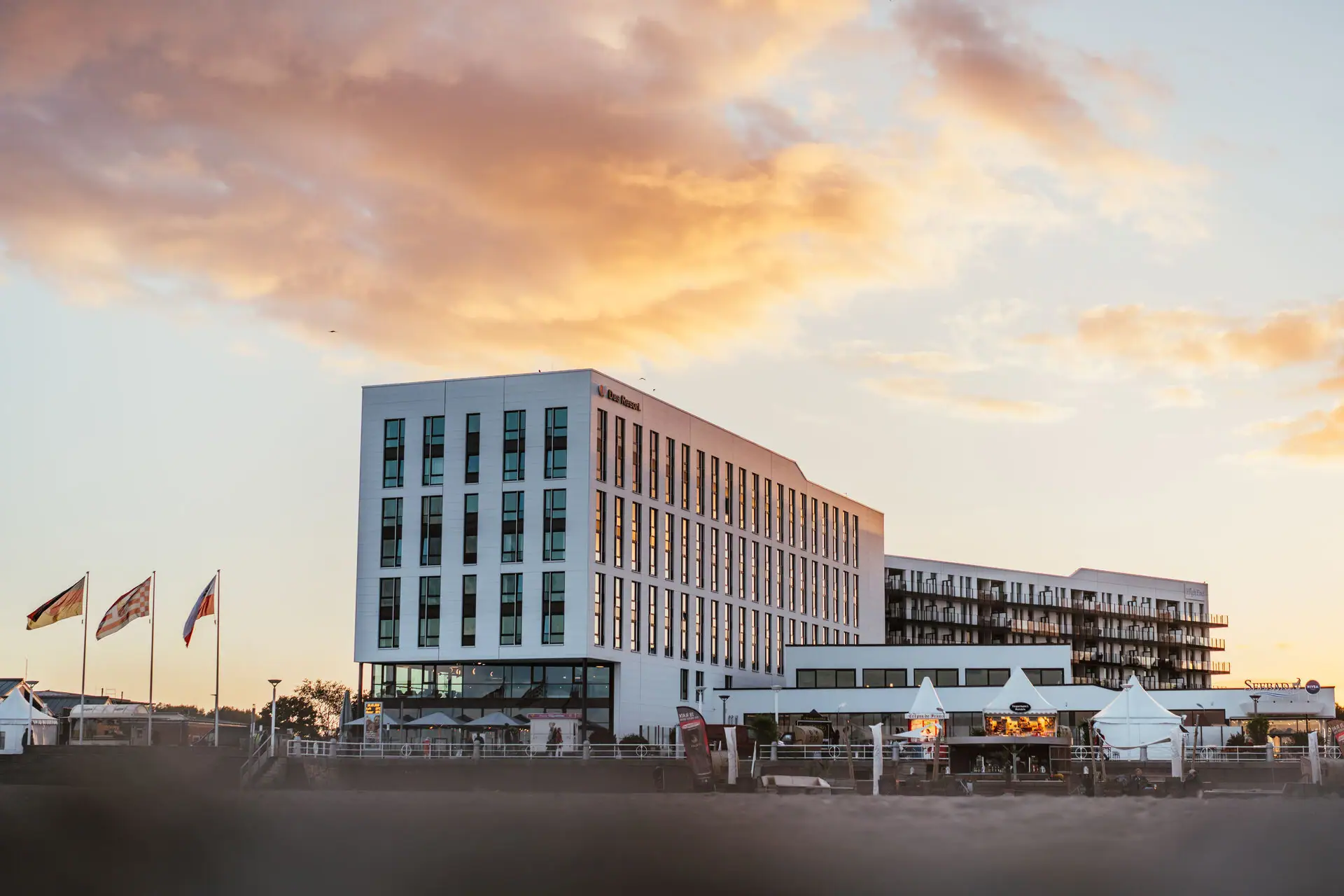 An exterior view of a white building with many windows on a beach at sunset.