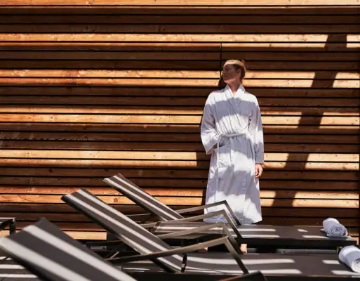 Wellness at the aja Bergresort Werfenweng A woman in a white bathrobe stands in front of a wooden wall.