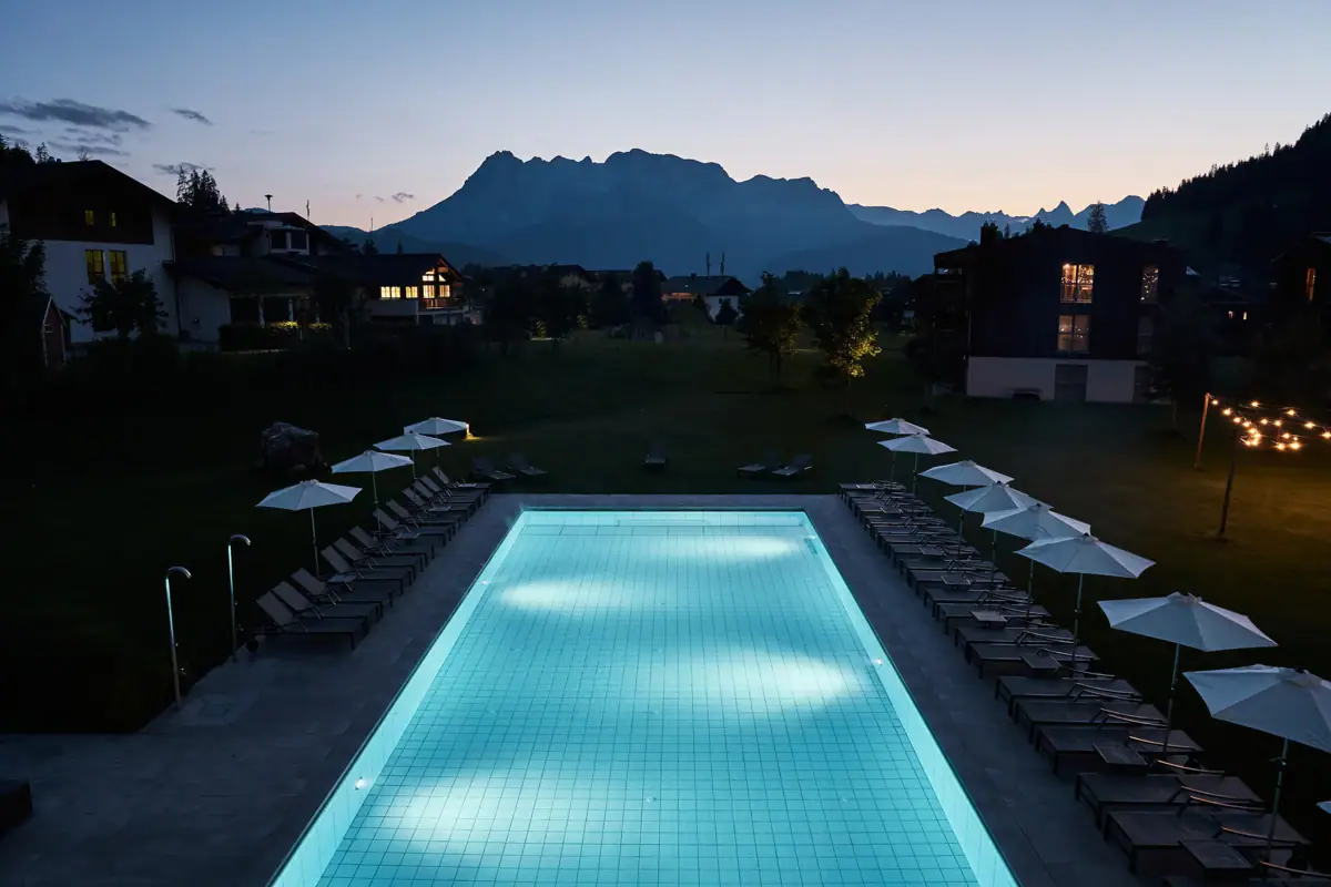 A pool with chairs and parasols right next to the hotel. The mountains on the horizon in the evening light.