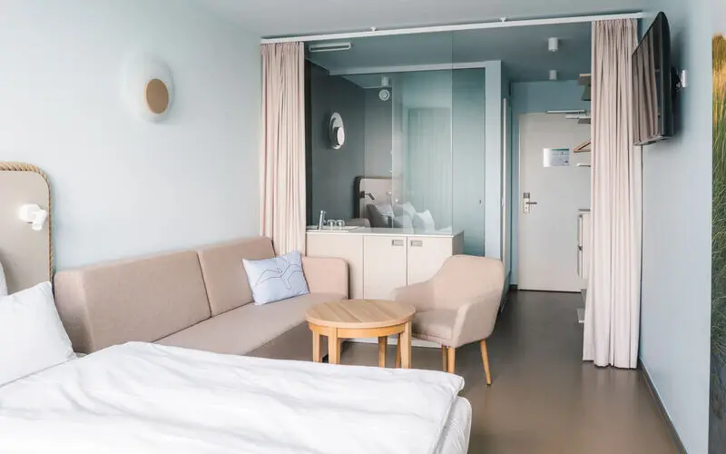 A cozy hotel room with pastel blue walls, a double bed, a small sitting area, and a modern bathroom partially separated by glass.