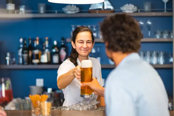 aja Bar Warnemünde A woman holds a glass of beer at the bar and hands it to a guest.
