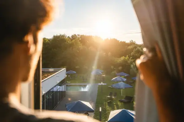 Room view A person looks at a pool with parasols and trees.