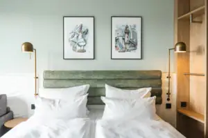 Rooms at the aja Warnemünde A double bed with white cushions and a green headboard, flanked by two lamps and an open wardrobe