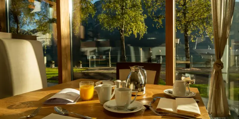 A breakfast table with a teapot and cups in front of a glass wall.