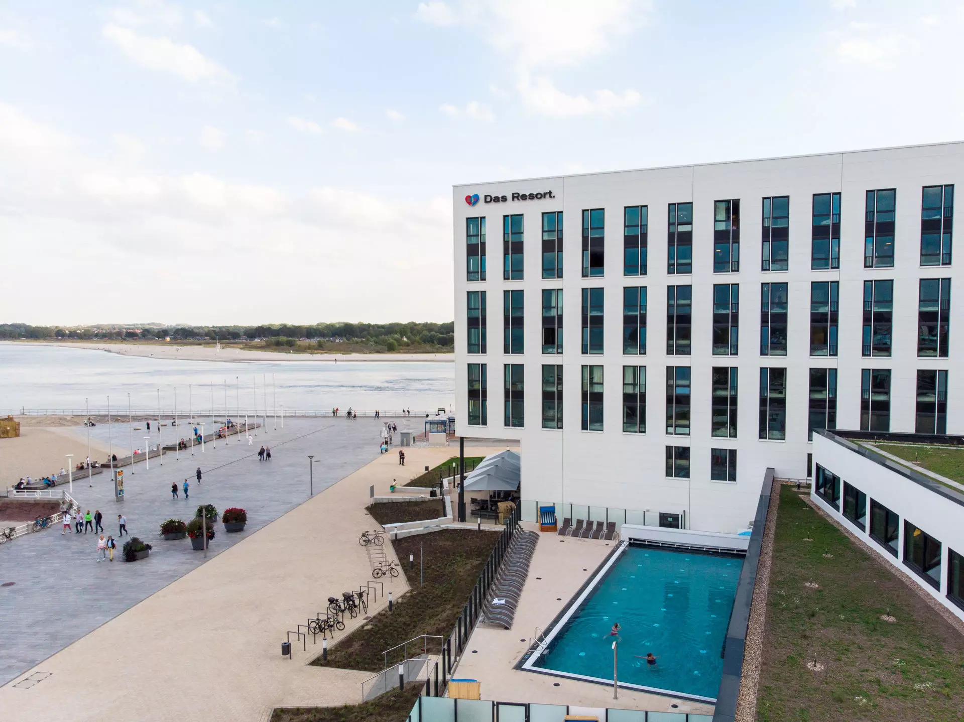 The modern facade of the hotel "Das Resort," featuring a swimming pool and spacious outdoor area, overlooking the beach and sea in the background.