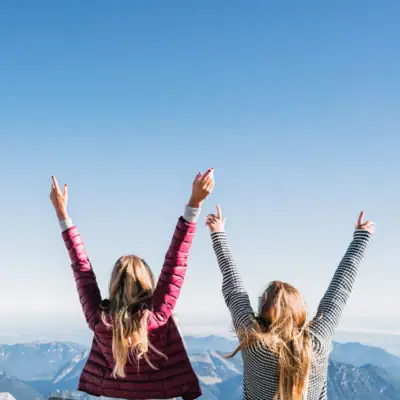 Two women are standing on a mountain with their arms raised.