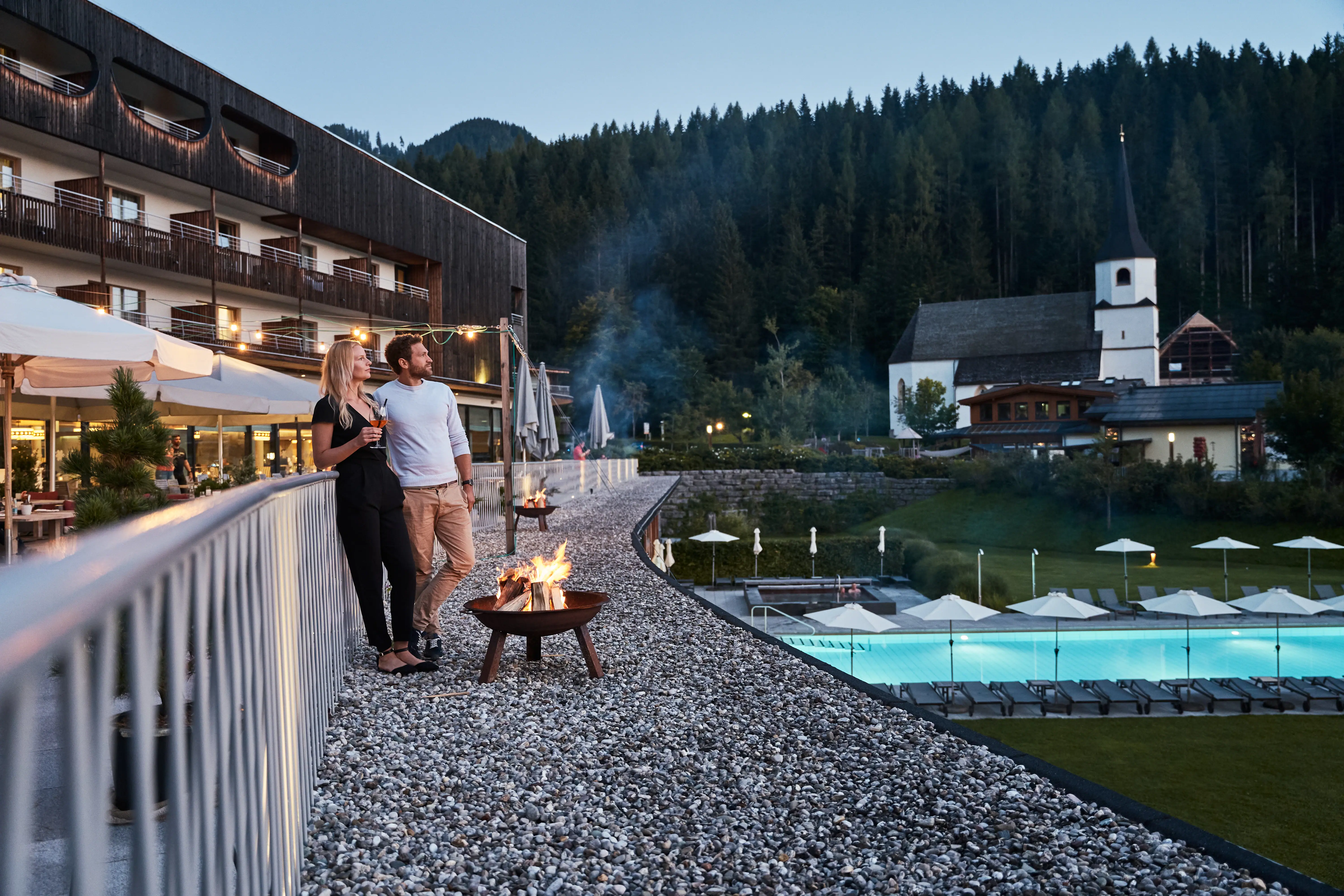 Couple sitting on the terrace of the hotel by a fire bowl in the evening, pool and church in the background.