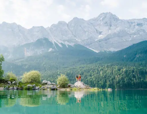 Two people are standing on a rock in front of a lake.