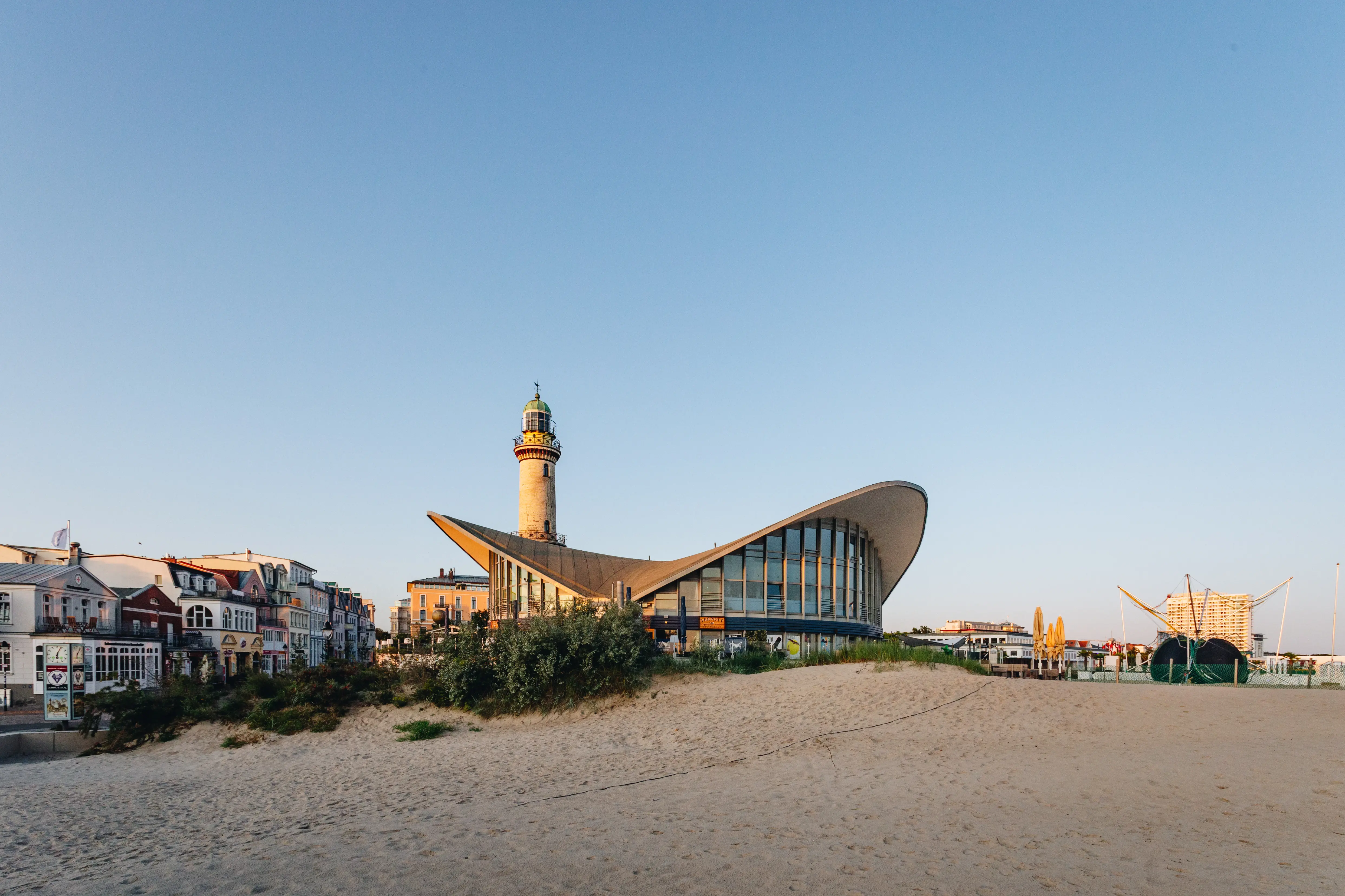 Iconic beach scene featuring the Warnemünde lighthouse and the distinctive curved architecture of the Teepott building, surrounded by sandy dunes and charming coastal houses under a clear blue sky.