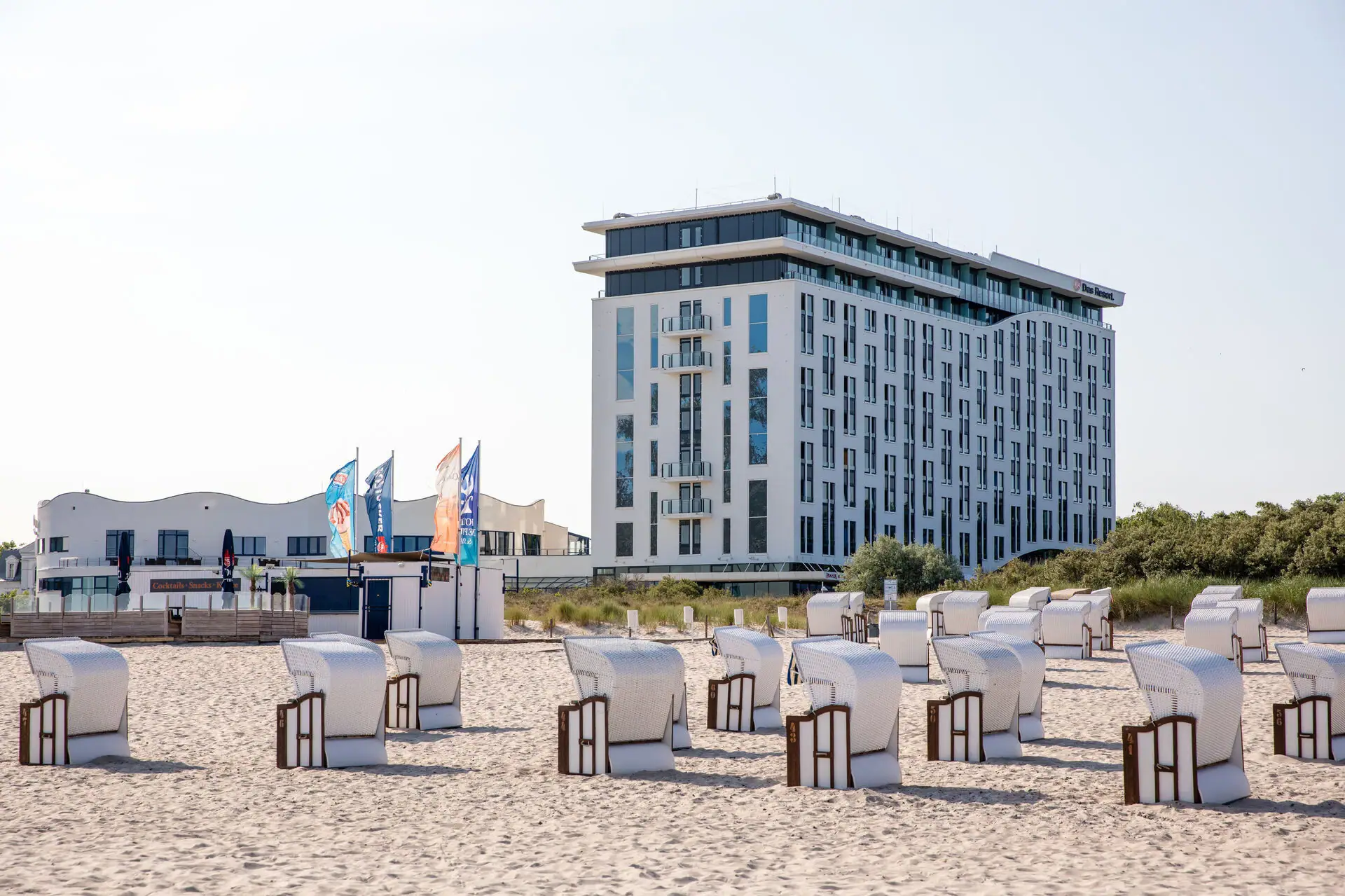 aja Warnemünde Beach chairs are lined up in front of a white building on the beach and the sun is shining.