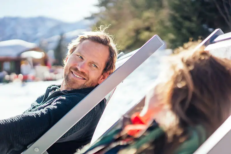 Man sitting on a deckchair outdoors, smiling, with a snowy landscape in the background.