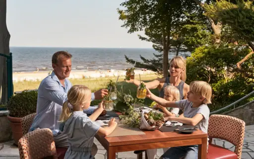 A family with three children sits on a terrace at a table with food on it and toasts with lemonade. The sea and the beach can be seen in the background.