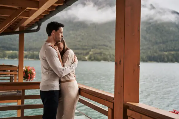 A man and a woman are embracing on a veranda overlooking a lake.