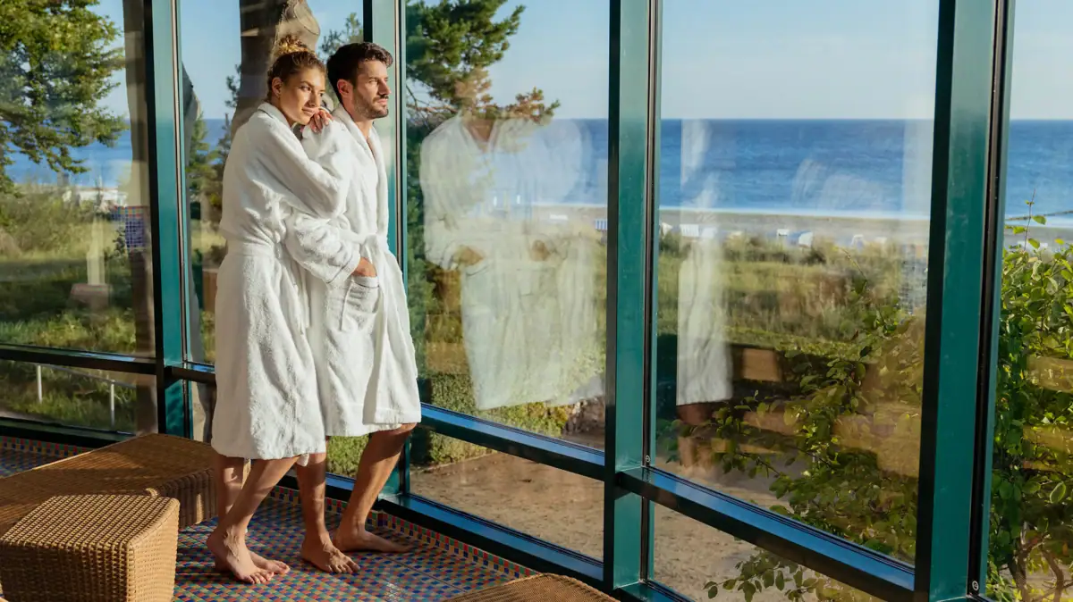 A man and woman in bathrobes standing in a room with large windows.