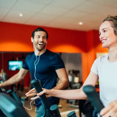 In a bright room with a wooden floor and white walls, two people are exercising and there are several cardio and strength machines lined up. The white wall at the back is illuminated by several lamps.