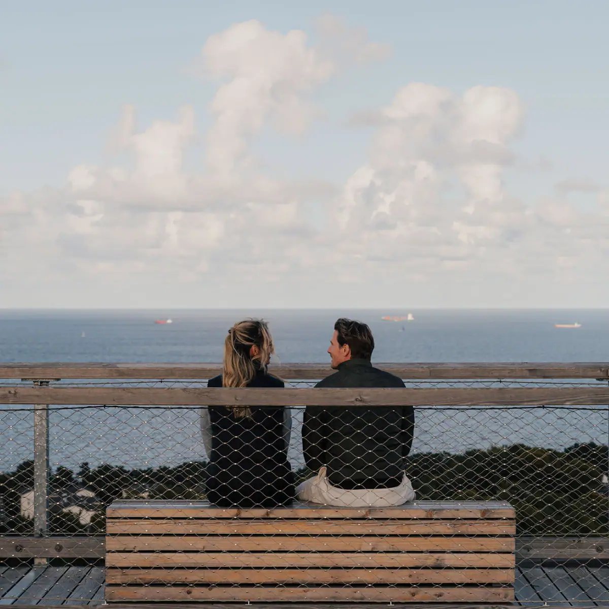 A man and a woman sit on a bench overlooking the sea.