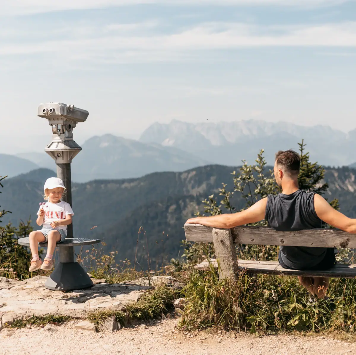 Father and daughter enjoy the view of the Alps from the Rosnerköpfl adventure mountain in Werfenweng. The girl is sitting on a lookout tower, the man is relaxing on a wooden bench with a view of the mountain panorama.