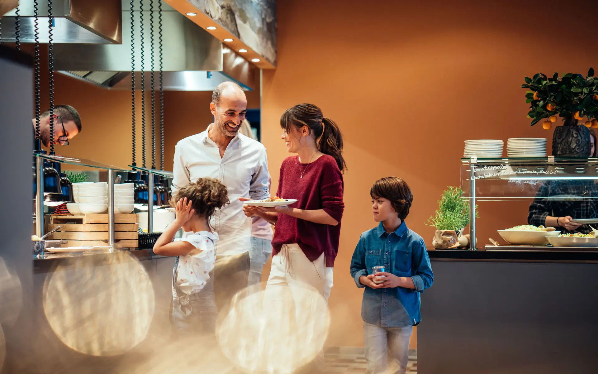 A family stands in a buffet area with plates in their hands and looks around. 