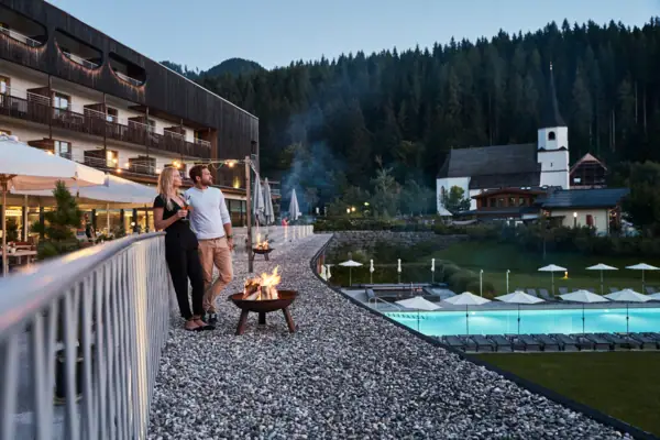 Couple sitting on the terrace of the hotel by a fire bowl in the evening, pool and church in the background.