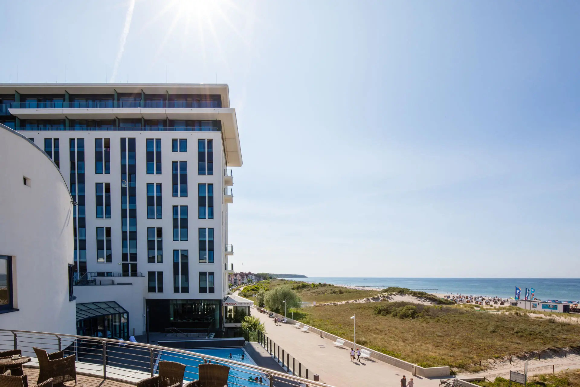 Modern beachfront hotel with a spacious terrace, outdoor pool, and a pathway leading to sandy dunes and the Baltic Sea under a bright, sunny sky.