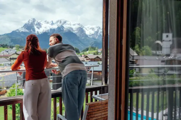 A man and a woman are standing on a balcony, looking out at the mountains.