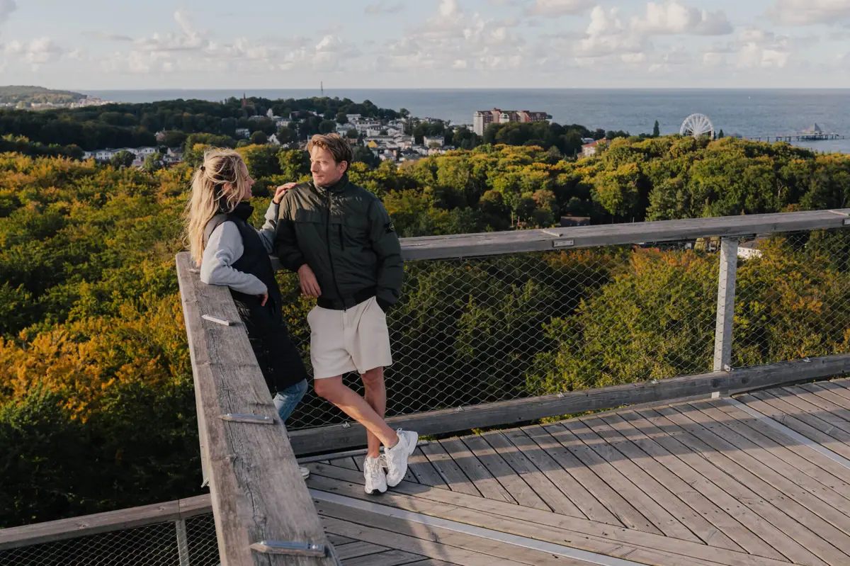 A man and a woman stand on a wooden deck overlooking a forest.