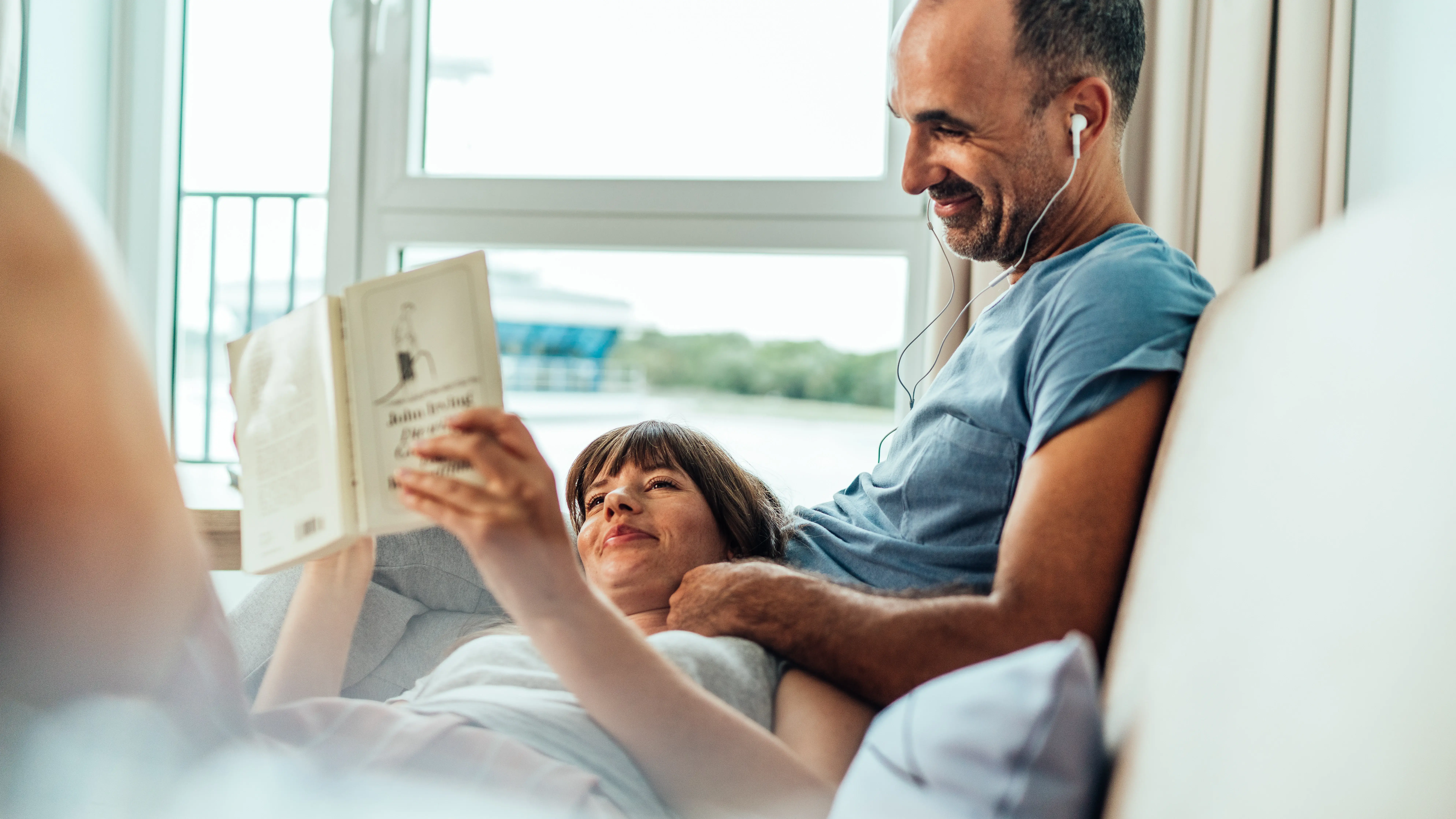 A cozy moment in a bright room with a couple relaxing on a couch; one person reading a book while the other listens to music with headphones, enjoying a peaceful atmosphere.