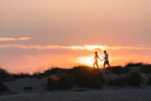 A couple holding hands on a sandy hill with a sunset in the background.