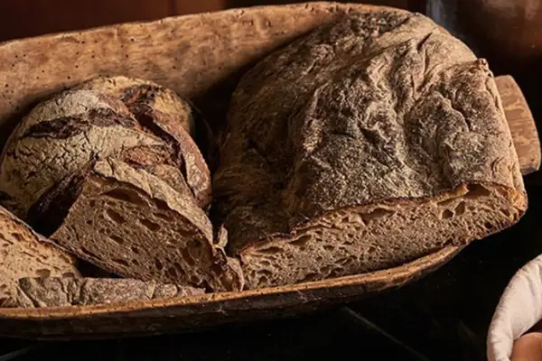 A loaf of bread and eggs in a basket next to a glass of milk.