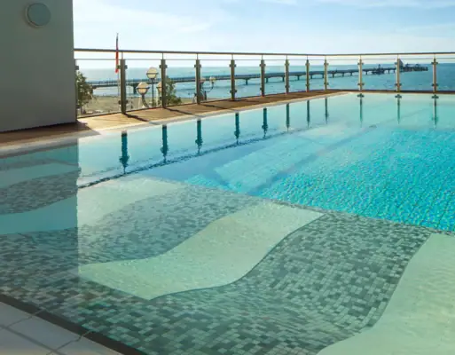 View over the calm waters of the Grömitzer Welle outdoor pool to the Baltic Sea in the background, which is separated from the outdoor area of the swimming pool by a glass fence.