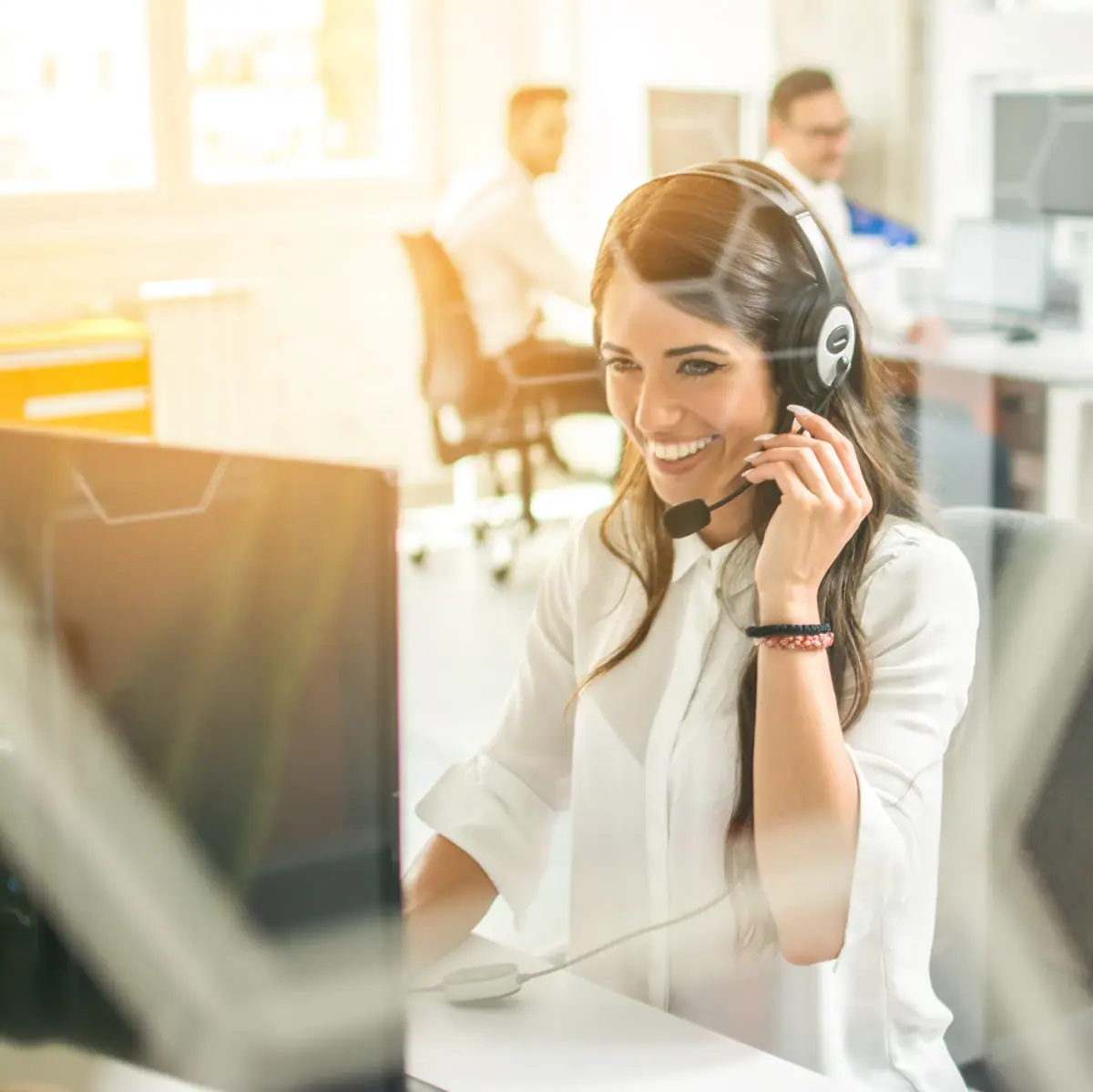 A smiling customer service employee wears a headset and works at a computer in a modern office with colleagues in the background.
