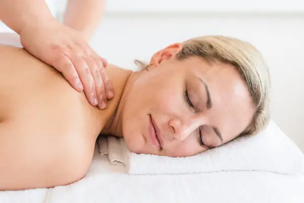 A woman lies on a white towel with her eyes closed and receives a neck massage.