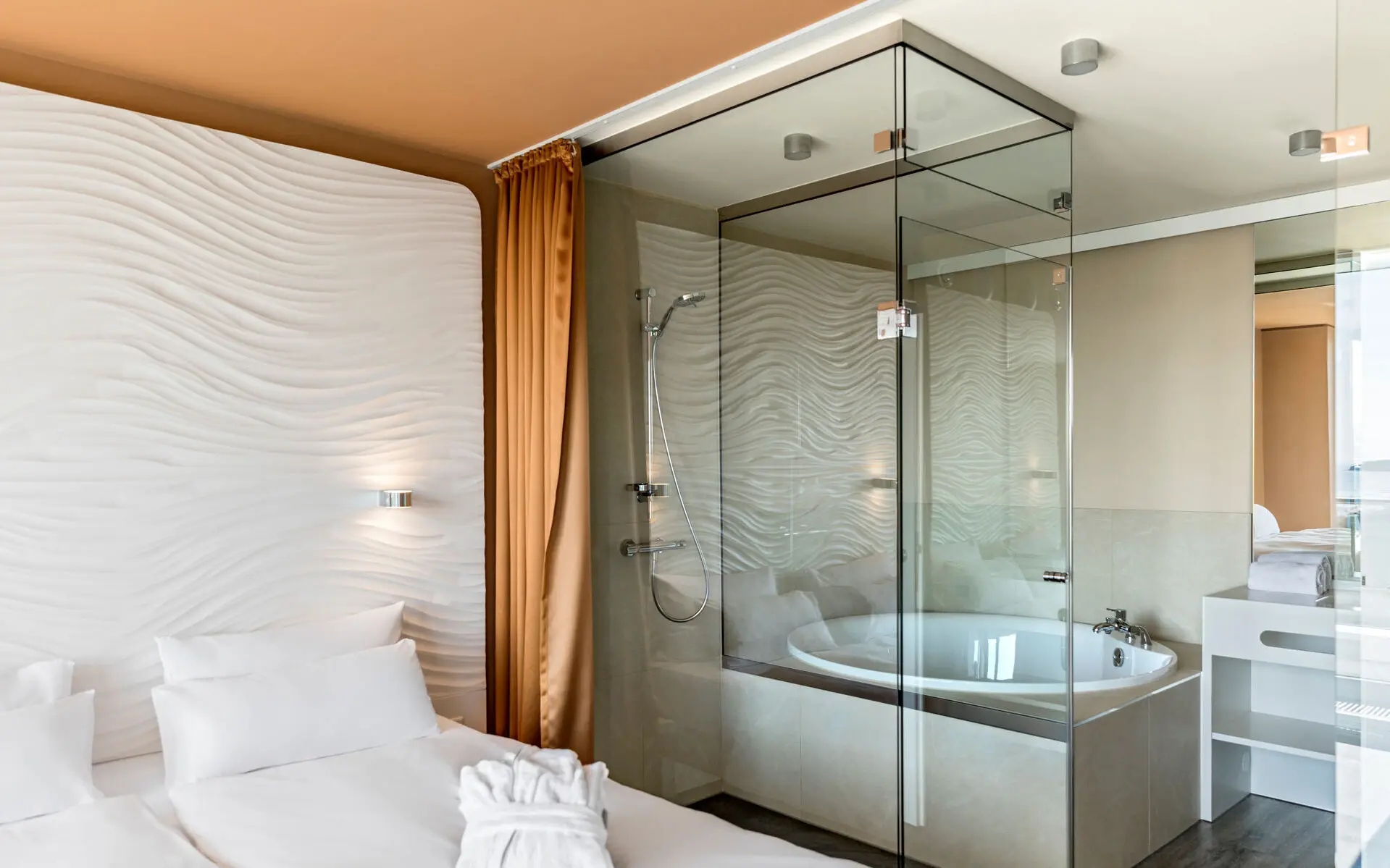 Modern hotel room with a comfortable double bed, stylish wave-patterned wall design, and an open-plan bathroom with glass partitions and a bathtub, creating a bright and luxurious atmosphere.