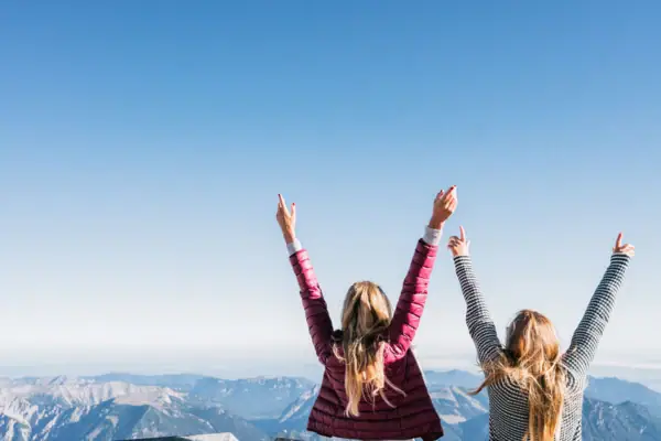 Two women are standing on a mountain with their arms raised.