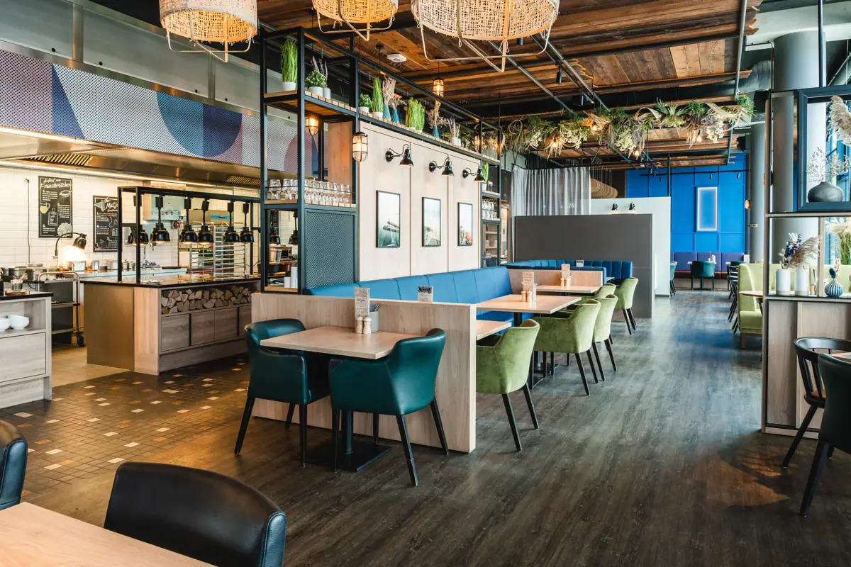 A restaurant with tables and chairs. The open buffet with lamps can be seen in the background.