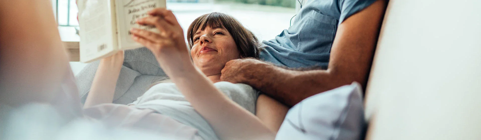 A couple is lying on a sofa. The man is listening to music and the woman is reading a book. 