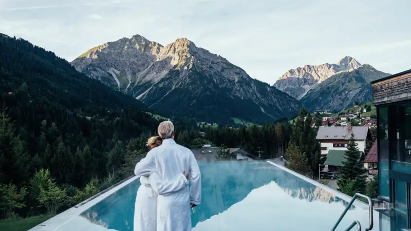 A man and a woman stand on a pool roof with mountains in the background.