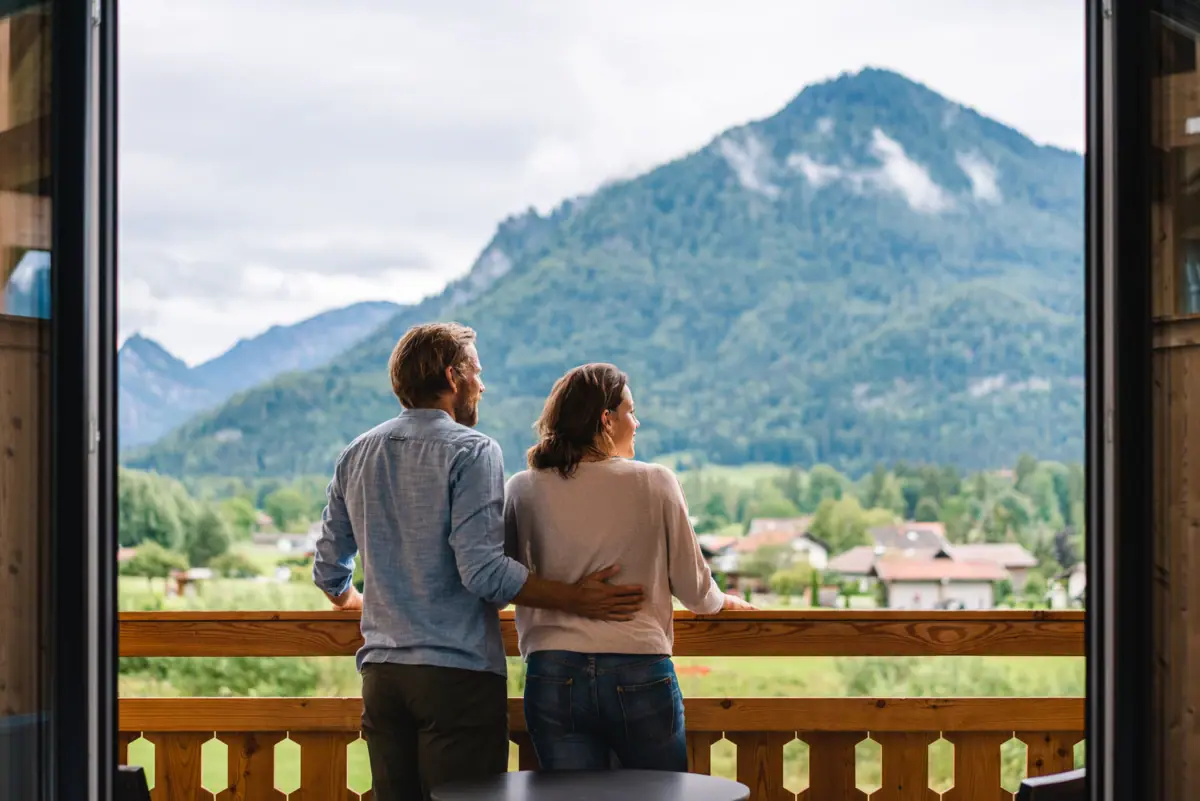 A man and a woman are standing on a balcony with a view of a mountain range.