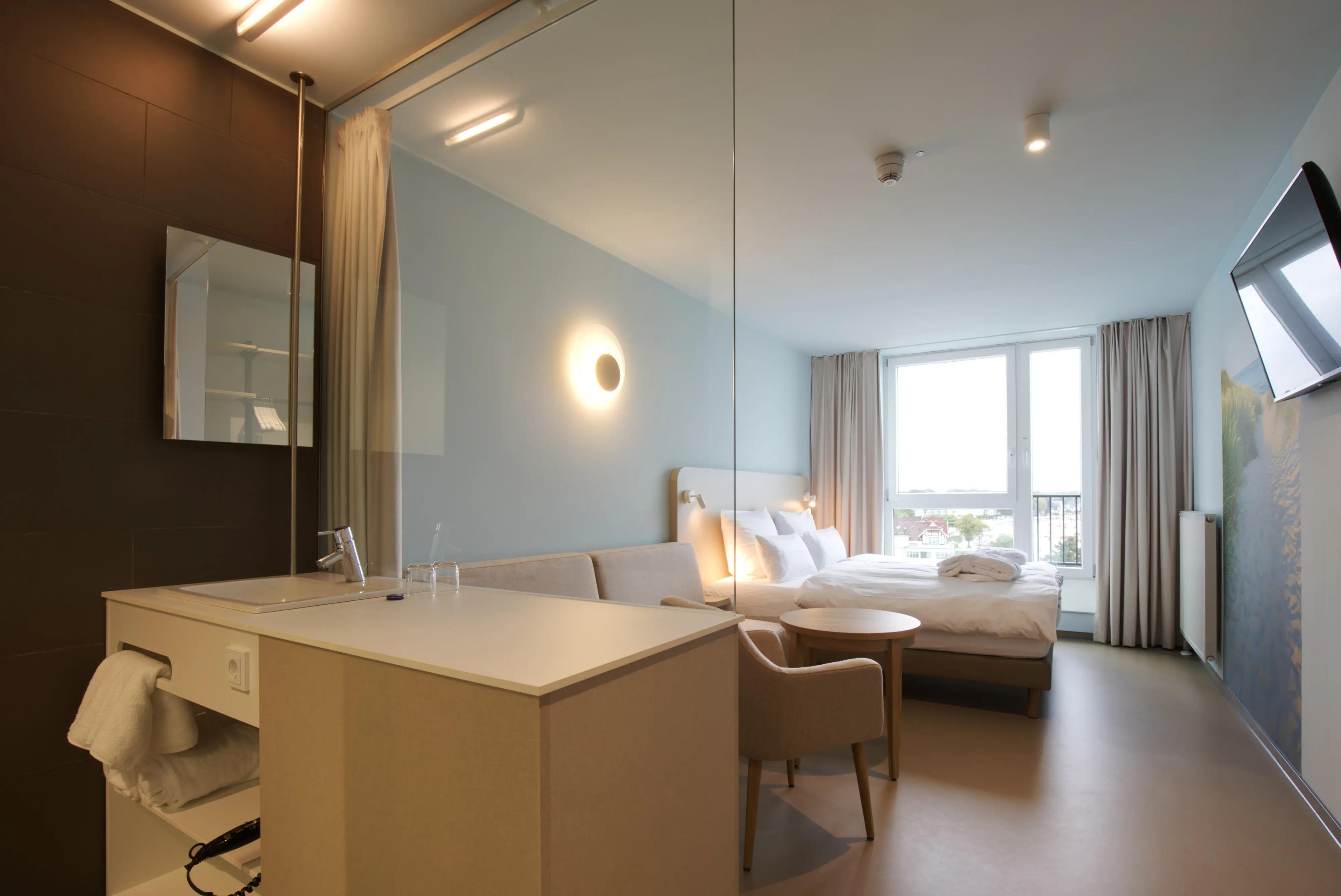 Modern hotel room with a minimalist design, featuring a comfortable bed, a seating area, and a partitioned open bathroom, illuminated by soft lighting and a large window with a view.